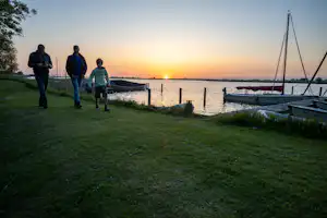 Camping am See bei Sonnenuntergang: zwei Personen am Ufer, Steg mit Booten und Ruderbooten im Wasser.