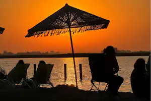 Sonnenschirm und Campingstühle am See beim Sonnenuntergang, goldene Spiegelung im Wasser und ruhige Uferlinie.