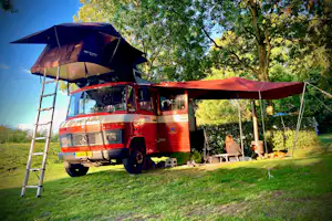 Roter Camper mit aufgestelltem Zelt und Sonnensegel auf dem Campingplatz, umgeben von Bäumen und Rasen.