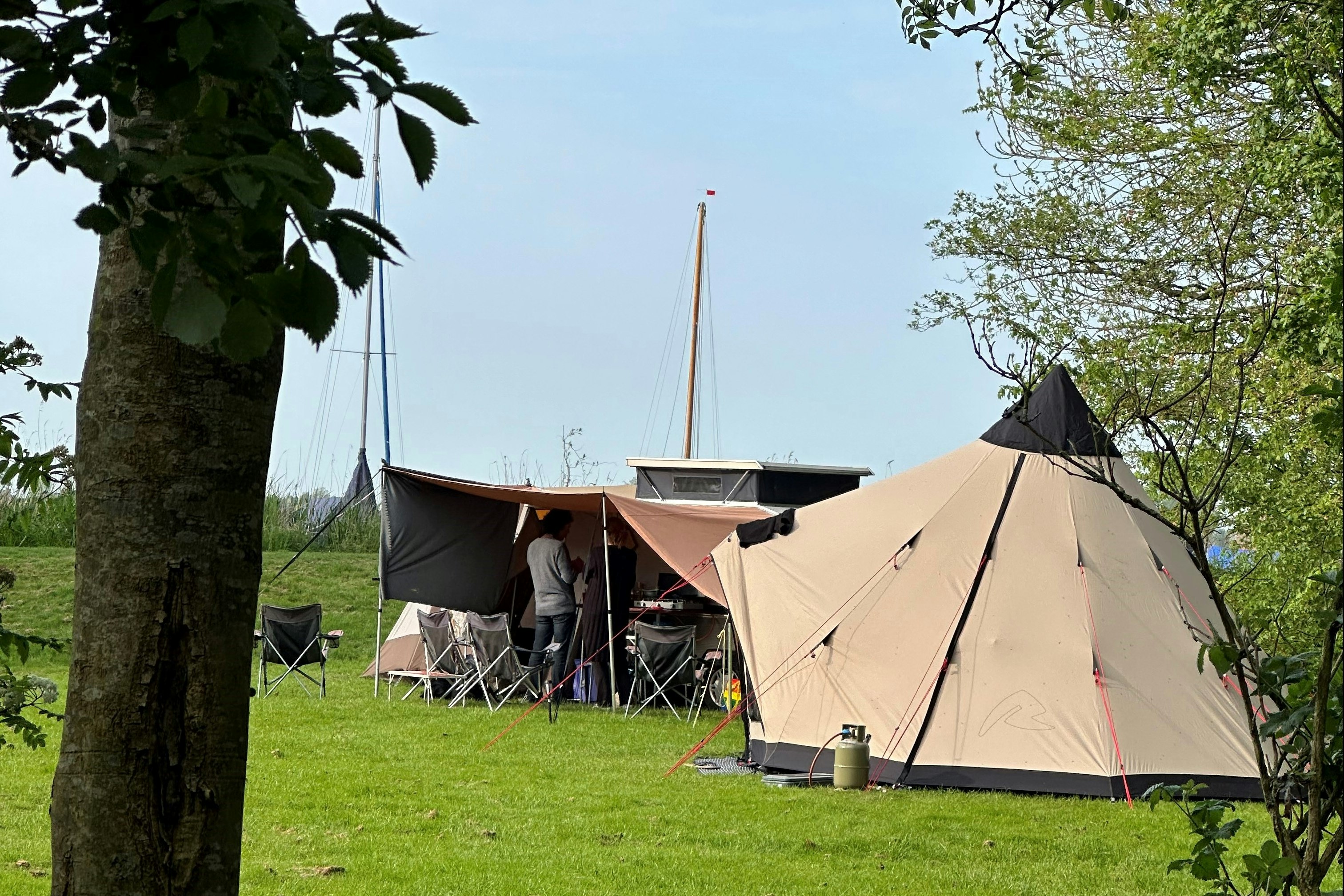 Campingzelt auf grünem Gras, daneben Klappstühle und Tisch, im Hintergrund Segelboot-Mast und weitere Zelte zwischen Bäumen.