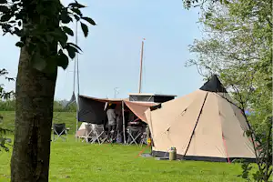 Campingzelt auf grünem Gras, daneben Klappstühle und Tisch, im Hintergrund Segelboot-Mast und weitere Zelte zwischen Bäumen.