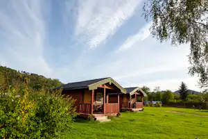 Holzferienhaus mit Veranda und Sitzbereich auf einer grünen Wiese, umgeben von Büschen und Bäumen, im Hintergrund Hügel.