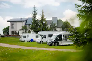 Mehrere Wohnmobile stehen auf einem grünen Rasen vor modernen Gebäuden, umgeben von Bäumen und kleinen Wegen.