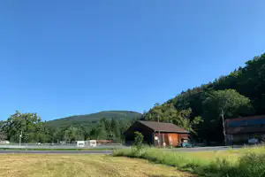 Holzgebäude auf einem Campingplatz mit Wiese, Zaun und Bäumen im Hintergrund vor grünen Hügeln.