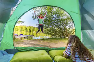 Kinder springen vor dem See, eingerahmt vom grünen Zelteingang mit Matratzen im Vordergrund und Steg am Ufer.