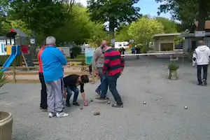 Petanque-Spiel auf dem Campingplatz: Boule-Kugeln auf dem Kiesplatz, Menschen am Bahnrand, Bäume und Gebäude im Hintergrund.