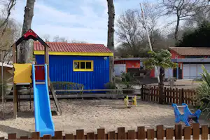Buntes Spielhaus mit Rutsche auf einem Campingplatz, umgeben von Bäumen und einem Kiesspielbereich.