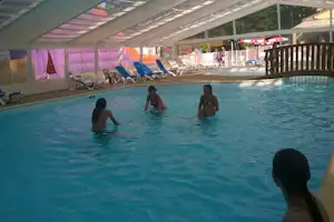 Innenpool mit mehreren Liegen unter einer großen Überdachung, im Hintergrund Zaun und Gebäude am Campingplatz.