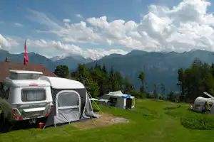 Campingplatz mit Wohnwagen und Vorzelt, grüner Wiese, Tisch und Stühlen sowie Blick auf Berge und Bäume.