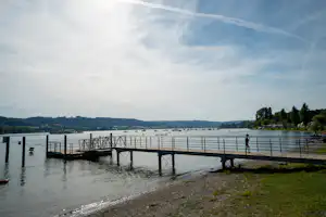 Steg am See mit Geländer, im Wasser liegen Segelboote, am Ufer Bäume und Hügel unter bewölktem Himmel.