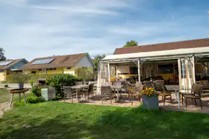 Camping-Terrasse mit überdachtem Sitzbereich, Gartenmöbeln und Tischen vor Bungalows, Hecken und Rasen im Hintergrund.