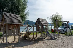 Holz-Spielplatz mit Rutsche und Klettergerüst am See, im Hintergrund Bäume, Wohnmobil und Zelt.