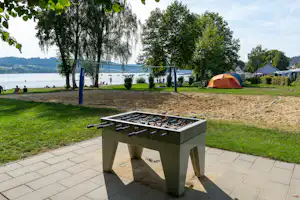 Kicker-Tisch auf der Terrasse am Sandstrand, im Hintergrund Volleyballnetz, Zelte und Bäume am Seeufer.