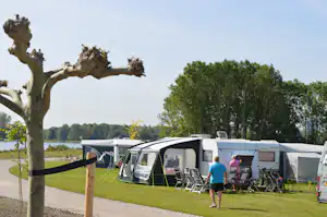 Campingplatz mit Wohnwagen und Mobilheimen auf einer Wiese, Stühle und Fahrräder am Stellplatz, Bäume im Hintergrund.