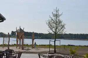 Spielplatz am Seeufer mit hölzernem Kletterturm, Seilen und Blick auf Wasser, Bäume und Strandweg.