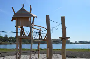 Spielplatz aus Naturholz mit Klettergerüst und Netzschaukel am See, im Hintergrund Bäume und blauer Himmel.