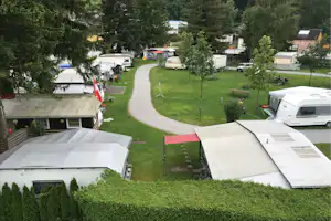 Campingplatz mit Wohnmobilen und Zelten auf Rasen, Kiesweg zwischen Stellplätzen, Bäume und Gartenmöbel im Hintergrund.