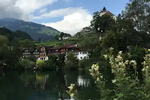 Blick auf ein Dorf am See mit Fachwerkhäusern, Bäumen und einem Kirchturm auf dem Hügel.