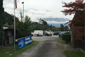 Camping-Einfahrt mit Parkplatz und Autos, im Hintergrund ein weißes Gebäude und Berge unter bewölktem Himmel.