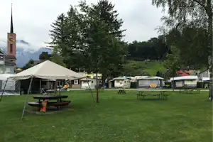 Campingplatz mit großen Zelten, Wohnwagen und Wohnmobilen auf einer grünen Wiese, im Hintergrund Bäume und Berge.