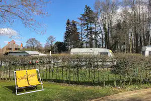 Wohnwagen mit Markise auf dem Campingplatz, im Hintergrund Bäume und ein Gebäude mit Schornstein.