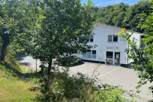 Weißes Haus auf dem Campingplatz, umgeben von Bäumen und Sträuchern, mit gepflasterter Terrasse und Eingangstür.