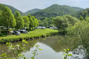 Campingplatz am Fluss mit Wohnwagen und Zelten auf der Wiese, umgeben von Bäumen und grünen Hügeln.