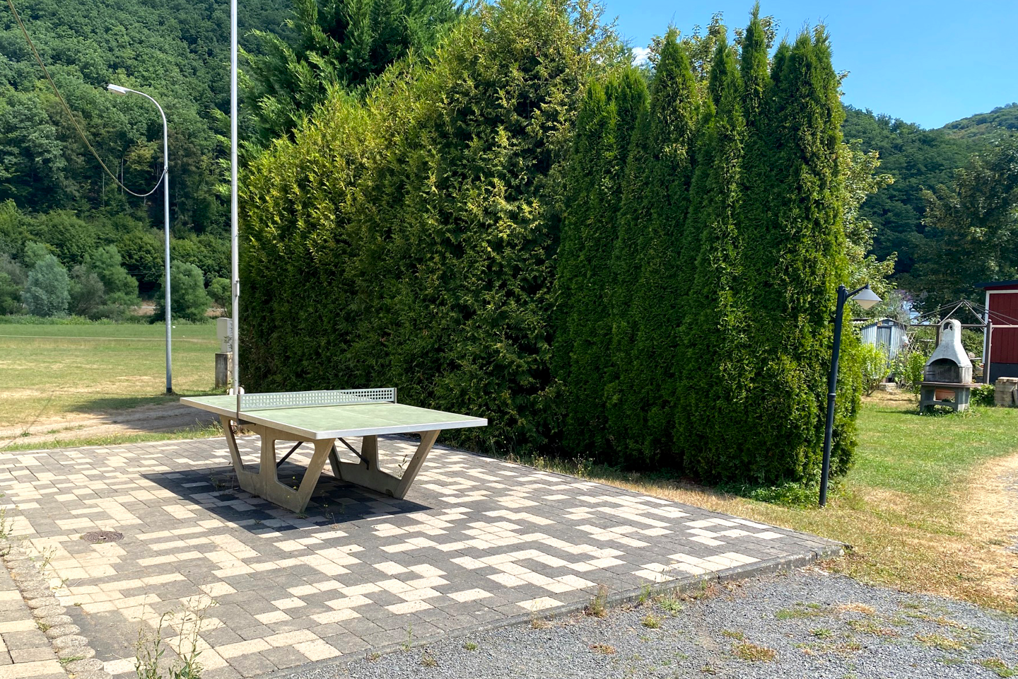 Picknicktisch auf gepflasterter Terrasse am Campingplatz, neben Hecke und Rasen, im Hintergrund Wald und Seeufer.