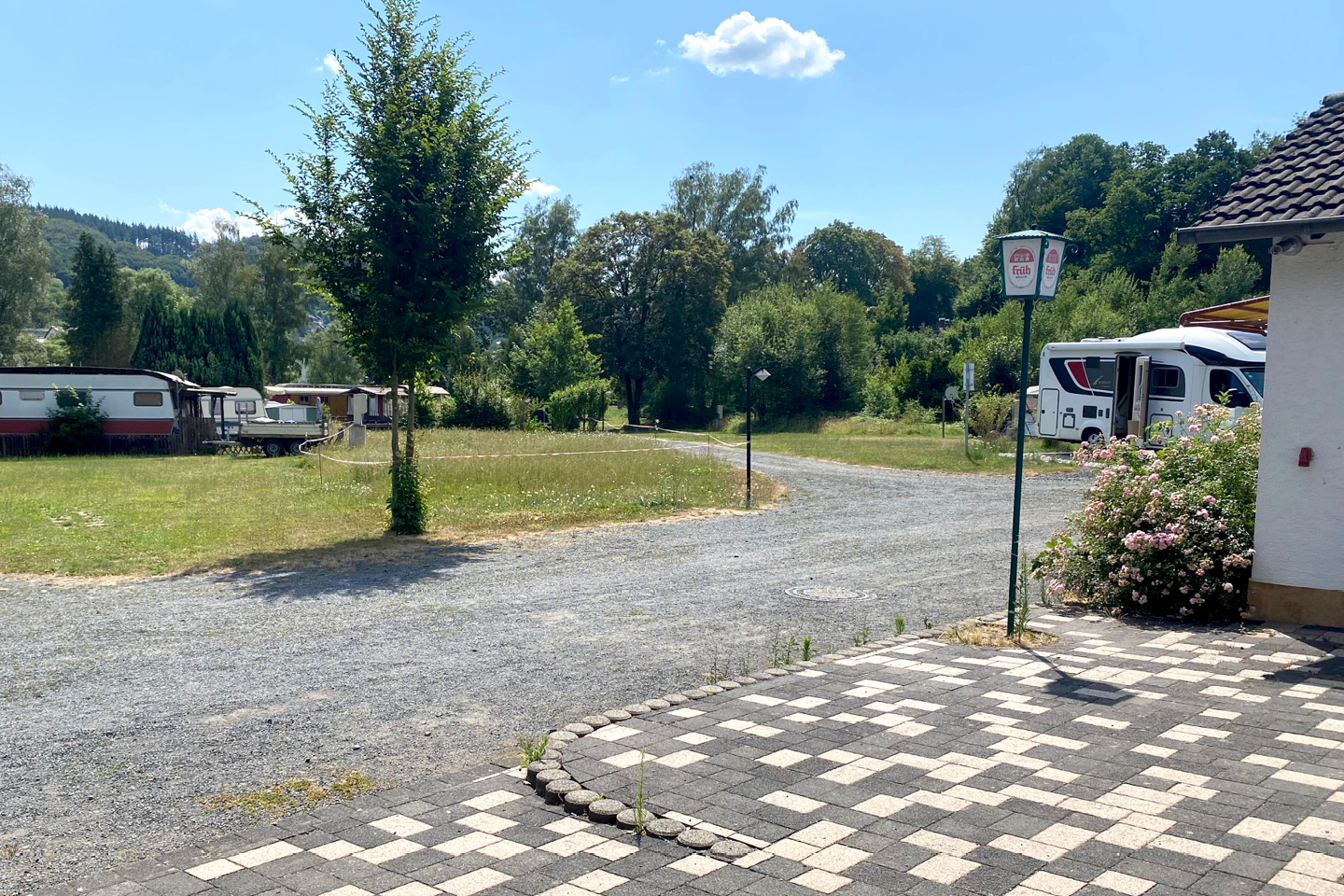 Stellplatz auf einem Campingplatz mit Kiesfläche, gepflasterter Terrasse, Wohnmobilen und Bäumen im Hintergrund.