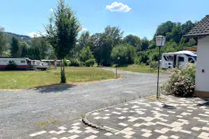 Stellplatz auf einem Campingplatz mit Kiesfläche, gepflasterter Terrasse, Wohnmobilen und Bäumen im Hintergrund.