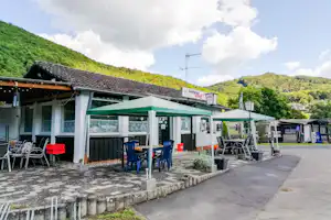 Campinggebäude mit Terrasse und grünen Markisen, Tischen und Stühlen, im Hintergrund Hügel und Bäume.