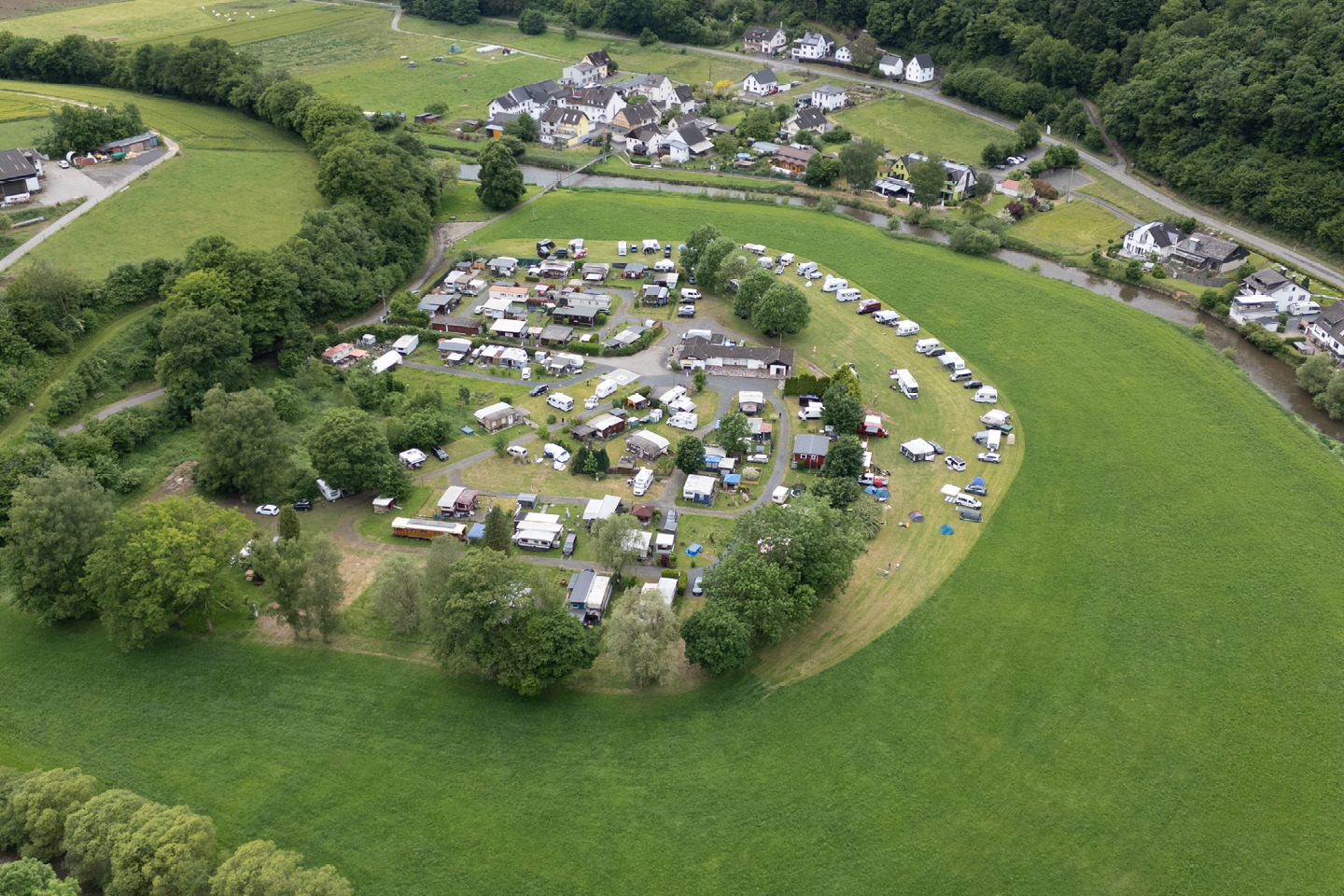 Luftaufnahme eines Campingplatzes mit vielen Stellplätzen, Wohnwagen und Zelten inmitten von grünen Wiesen und Bäumen.