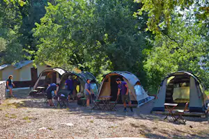 Mehrere Zelte auf einem Campingplatz, mit Holzstühlen und Tischen vor den Eingängen, umgeben von Bäumen im Grünen.