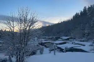 Verschneiter Campingplatz mit Wohnwagen und Zelten, davor Schnee auf Bäumen und Hecken bei blauem Abendhimmel.