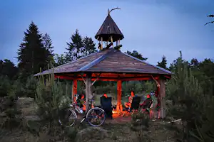 Camping-Pavillon mit Satteldach, davor Fahrräder und Sitzgelegenheiten, umgeben von Bäumen und Sträuchern.