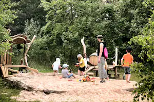 Spielende Kinder auf einem Sandbereich am Campingplatz im Wald, umgeben von Bäumen und Holzspielgeräten.