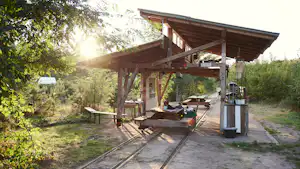 Holzüberdachung mit Grill und Tisch auf einem Campingplatz, umgeben von Bäumen und einem gepflasterten Weg.