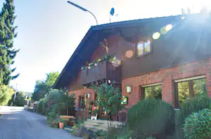 Ferienhaus mit Ziegelwand, Holzfassade und Balkon, Fenster und Lampen am Eingang, umgeben von Bäumen und Sträuchern.