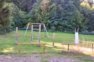 Spielgerät aus Holz auf grünem Campingplatz, im Hintergrund dicht bewachsene Bäume und Wiese.