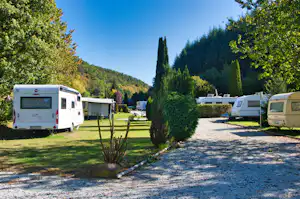 Wohnmobile auf einem Campingplatz-Stellplatz, gepflasterter Weg, Hecken und Bäume vor grünen Hügeln im Hintergrund.