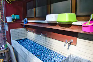 Außenwaschbecken mit blau-weißen Kacheln, Holzablage und Wasserhähnen, daneben bunte Eimer und Schüsseln.