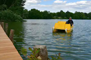 Gelbes Ruderboot auf dem See neben einem Holzsteg, im Hintergrund Bäume und ein Mann im Boot.