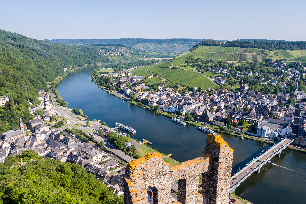 Blick auf einen Fluss mit Stadt und Uferpromenade, im Vordergrund eine alte Mauer und Türme.