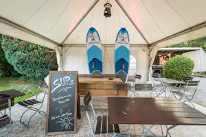 Unter einem großen Campingzelt stehen SUP-Boards und ein Holzschrank, daneben Stühle und ein Tafel-Schild.