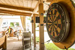 Dartscheibe mit Pfeilen im Zelt, Holzbalken und Sitzbereich mit Tischen und Stühlen im Hintergrund.