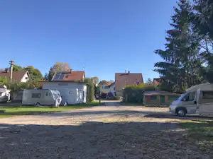 Stellplatz auf dem Campingplatz mit Wohnwagen und Wohnmobil, Kiesfläche, Bäumen und Häusern im Hintergrund.