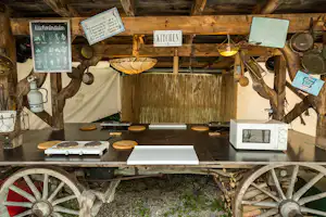 Rustikale Campingküche mit Holzbalken, Schildern „Küchenkräuter“ und „Kitchen“, Kochplatten, Pfannen und Hängelampen.