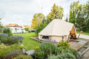 Campingzelt auf einem Stellplatz im grünen Garten, mit Holzpodest, Steinen und Büschen, im Hintergrund Autos und Wohngebäude.
