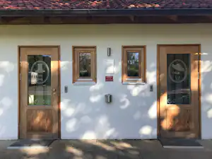 Weiße Hauswand mit zwei Holztüren und Fenstern, davor ein gepflasterter Weg und Schatten von Bäumen.