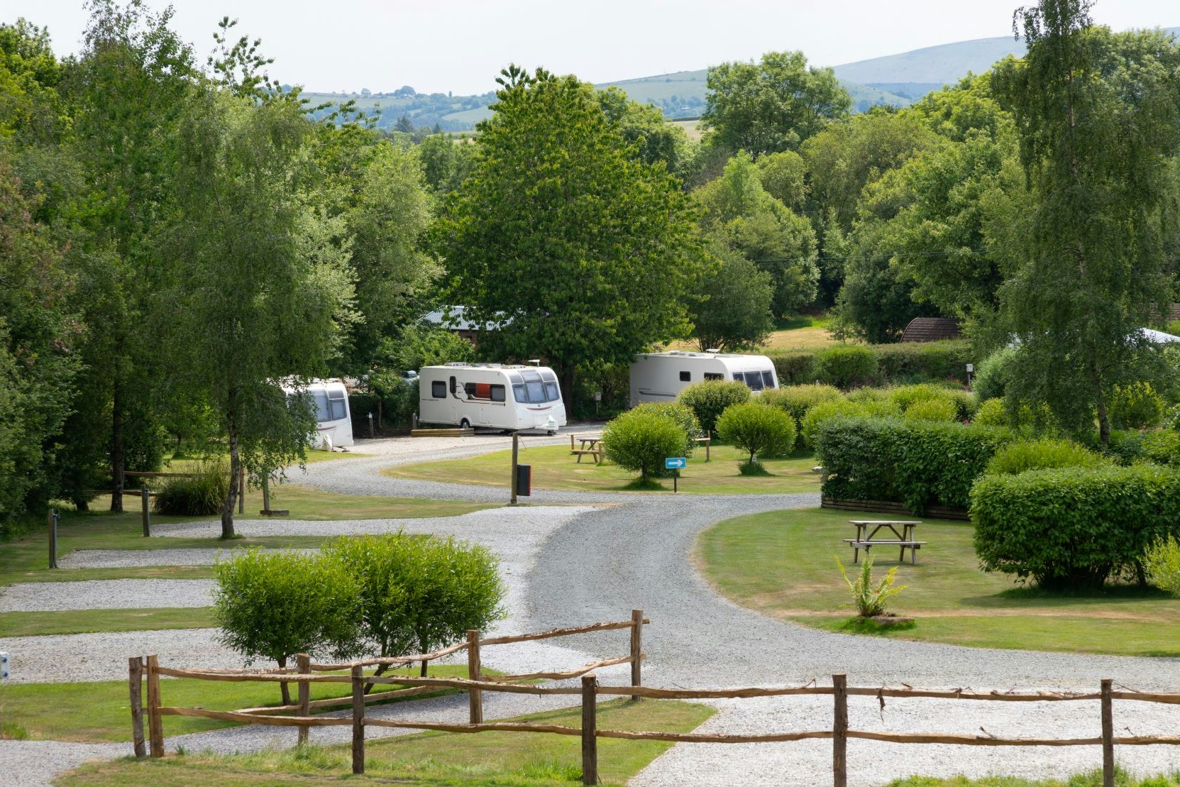 Wohnwagen auf einem Campingplatz, gepflasterter Weg und Hecken zwischen grünen Bäumen, im Hintergrund Hügel und Tal.